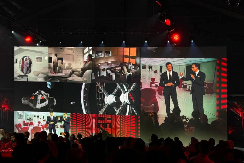 A large event screen displaying a variety of images including astronauts, spacecraft interiors, and two men conversing beside a Ferrari 12Cilindri sports car, viewed by an audience under red lighting.