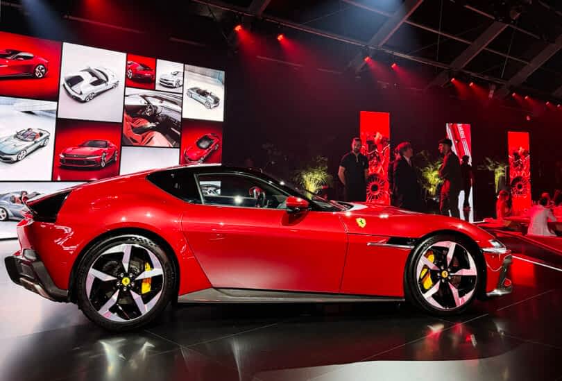 Red Ferrari 12Cilindri sports car on display at an auto show with people and promotional images in the background.