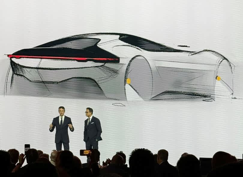 Two men presenting a Ferrari 12Cilindri sports car sketch on a large screen at an automotive event.