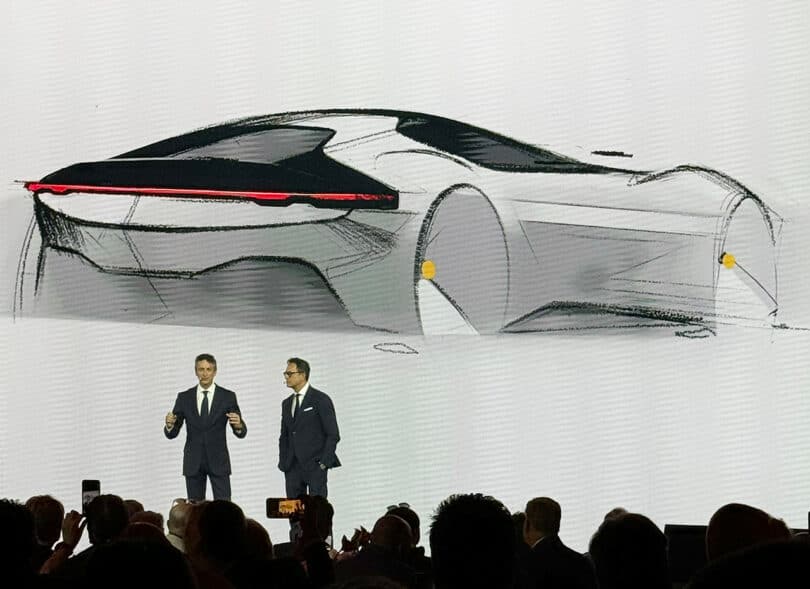 Two men presenting a Ferrari 12Cilindri sports car sketch on a large screen at an automotive event.