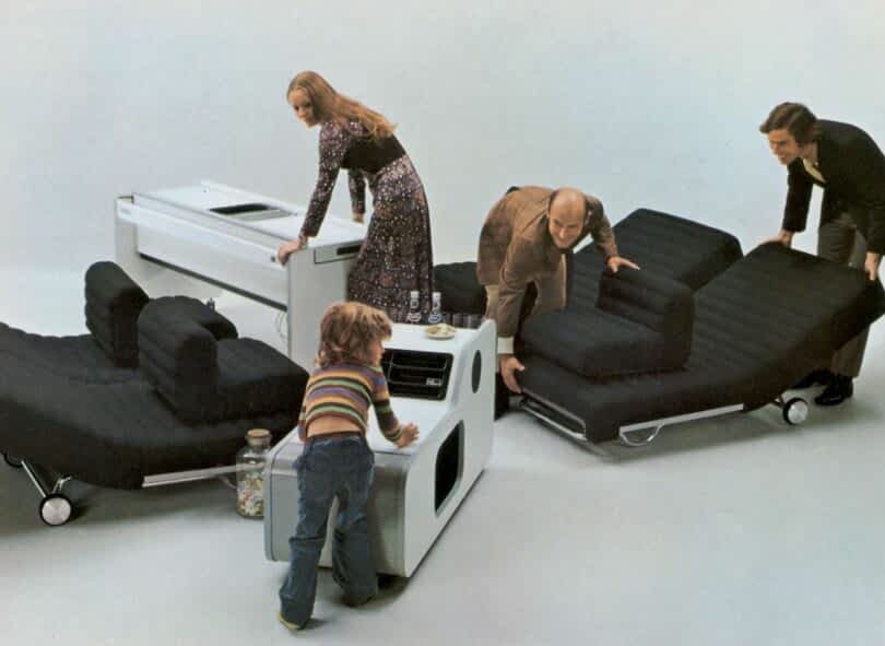 Three adults and a child interact with modular black furniture arranged in various configurations on a plain backdrop