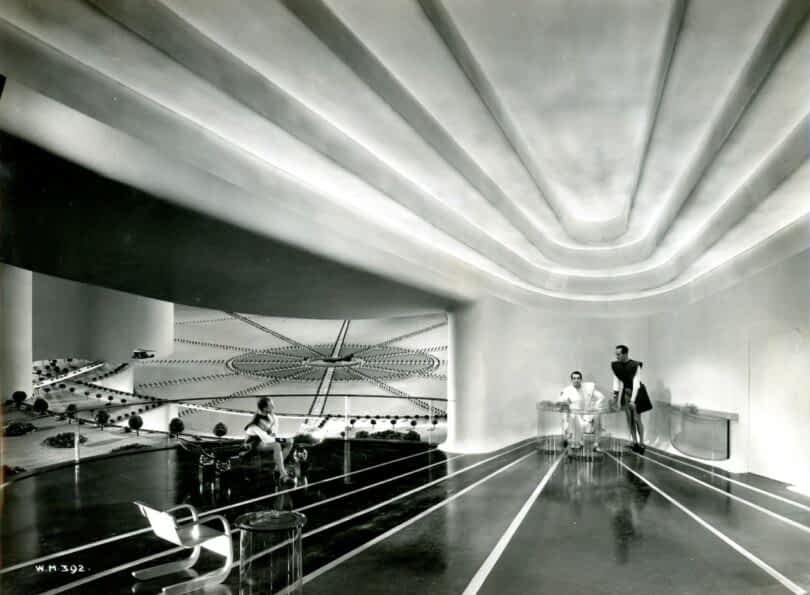 A futuristic room with curved ceiling beams, abstract wall art, and three people interacting – one seated on the left, one standing center-right, and one seated at a transparent desk in the center