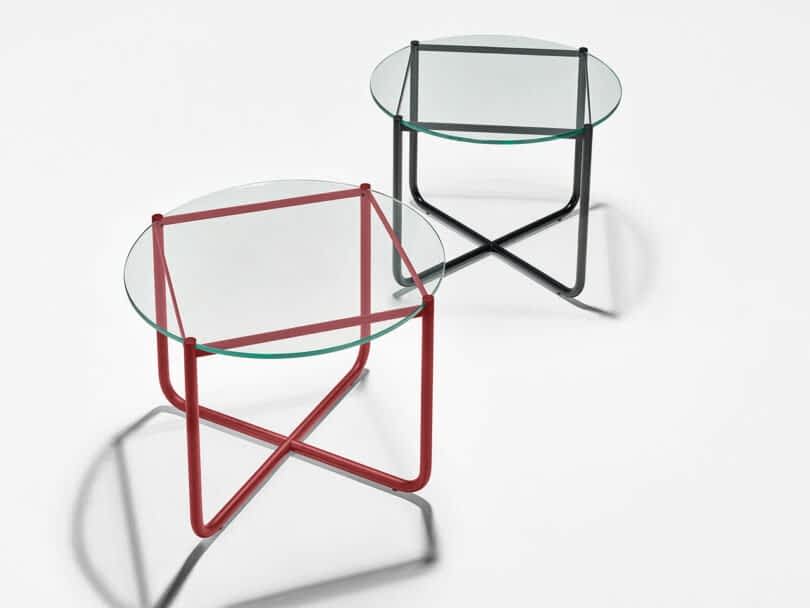 Two modern glass-top side tables with intersecting tubular metal legs, one in red and the other in black, placed on a white surface.