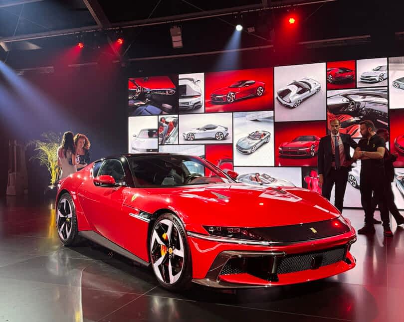 A Ferrari 12Cilindri on display at an automotive event, surrounded by people and large screens showing car designs.
