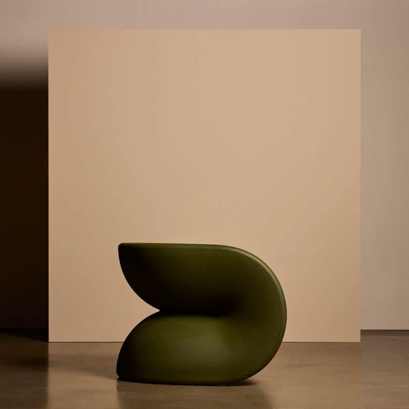 A modern, sculptural chair with smooth, curved lines in dark green, placed in front of a beige backdrop.
