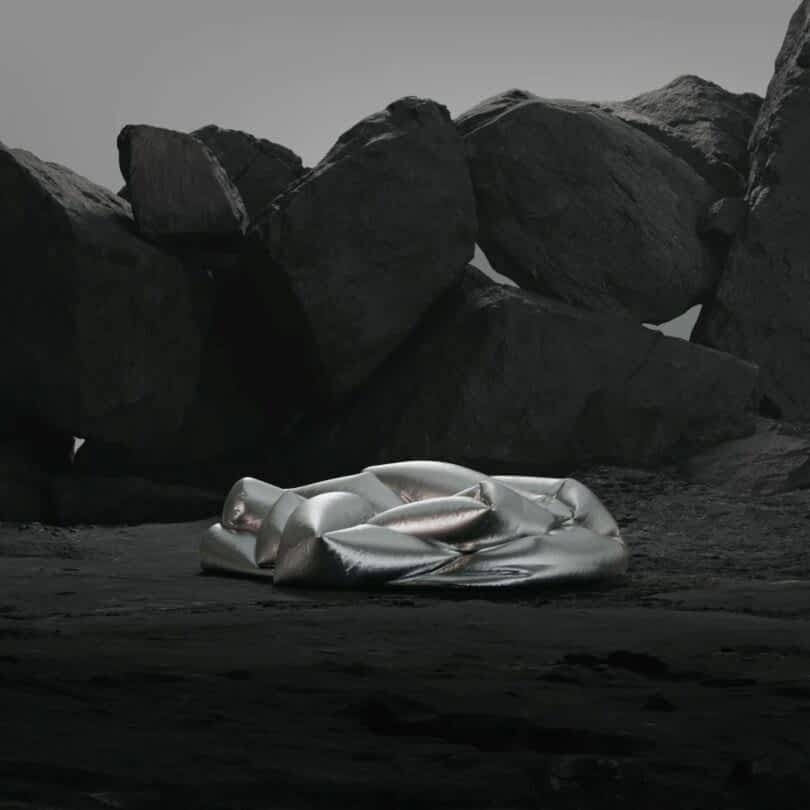 A metallic, inflated sofa lies on a rocky terrain with large boulders in the background under a gray sky