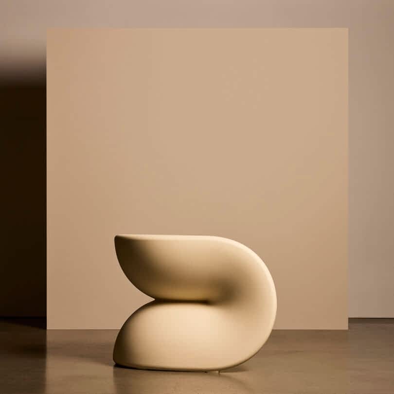 A modern, sculptural chair with smooth, curved lines in cream, placed in front of a beige backdrop.