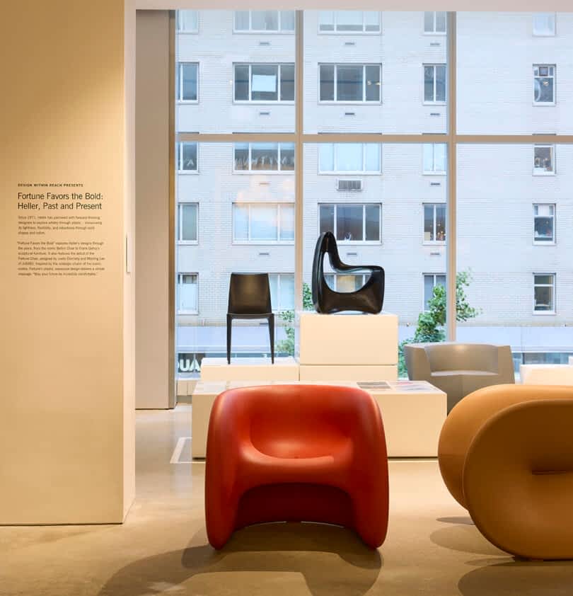 An exhibition room features modern designer chairs, including red, brown, and white pieces, displayed on white podiums in front of large windows with a cityscape view.