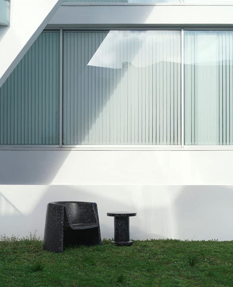 A black lounge chair and matching table sit on grass in front of a building with large windows featuring vertical blinds.