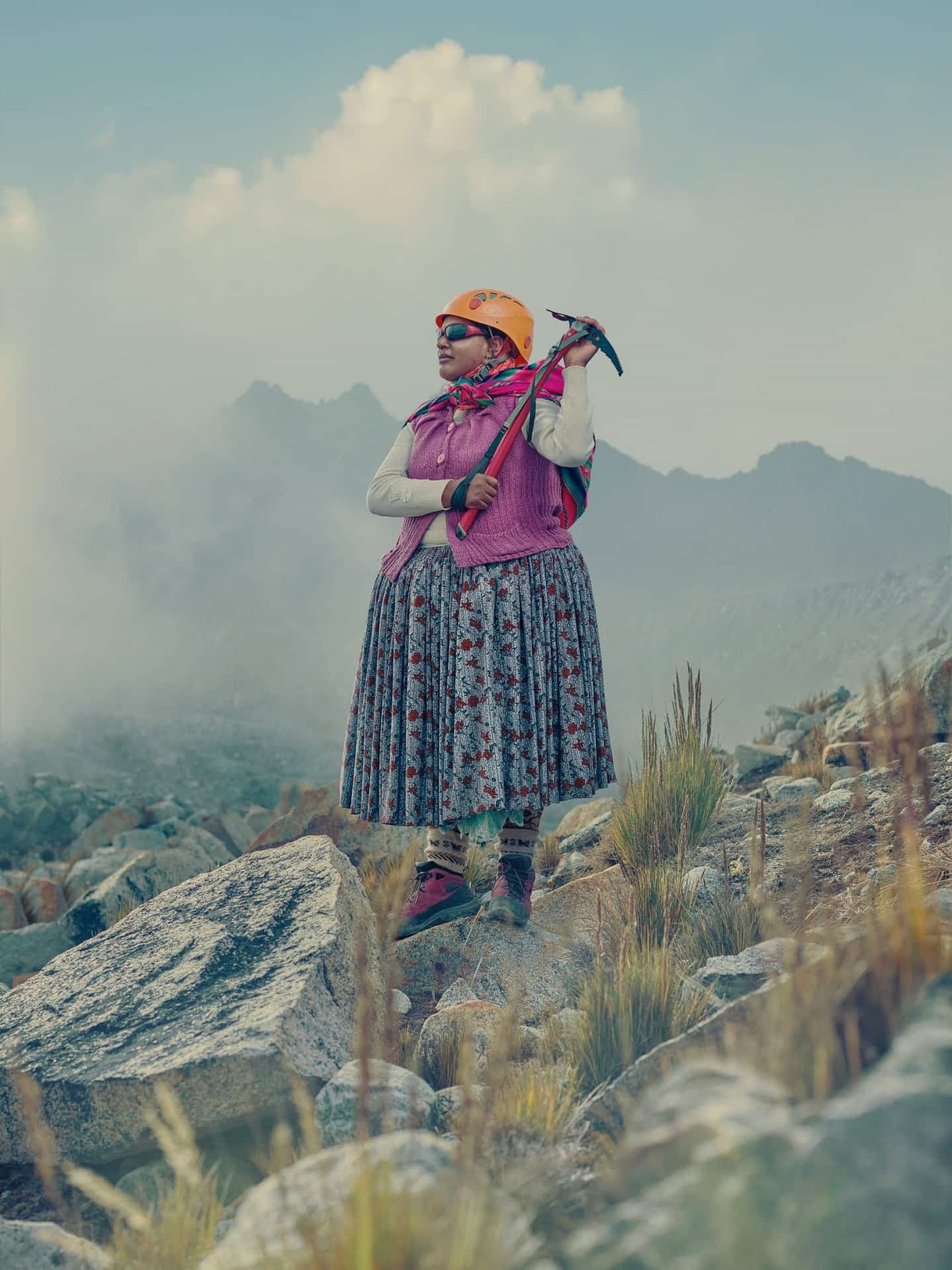 an Bolivian Indigenous Aymara woman stands on a mountain, wearing an orange helmet and colorful skirt, holding an ice pick