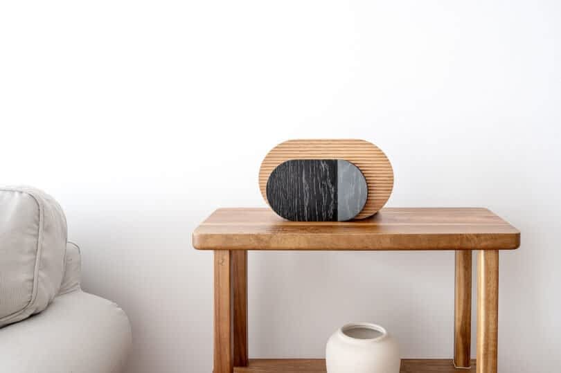 A wooden side table with decorative objects: a modern, oval-shaped wooden and black lamp, and a small, light-colored ceramic vase. Part of a light gray couch is visible on the left.