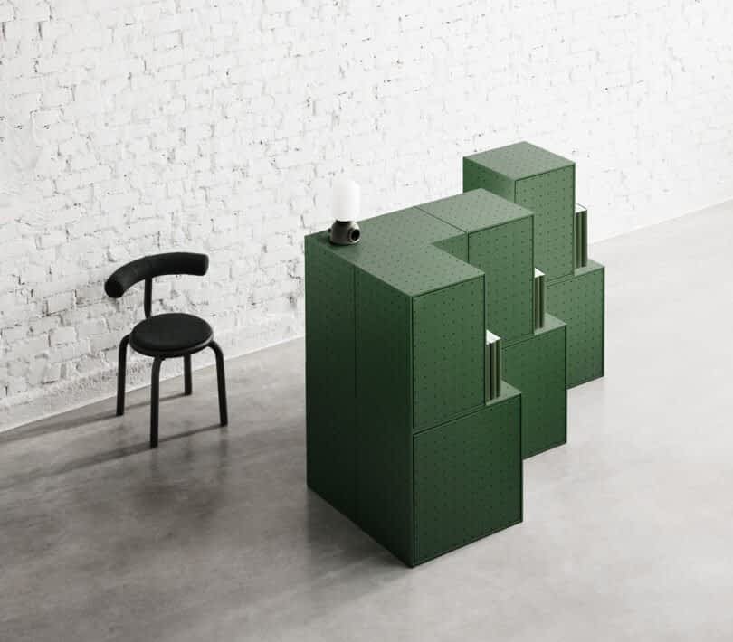 A modular, green storage unit from the String Furniture X FUWL Center collection is composed of blocks and is placed near a minimalist black chair in front of a white brick wall in a modern, sparsely furnished workspace.