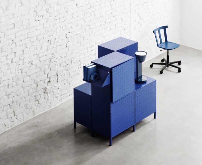 Blue modular cabinets and an office chair on wheels enhance this workspace furniture in a room with a white brick wall and gray floor, featuring the sleek design of String Furniture X FUWL Center.