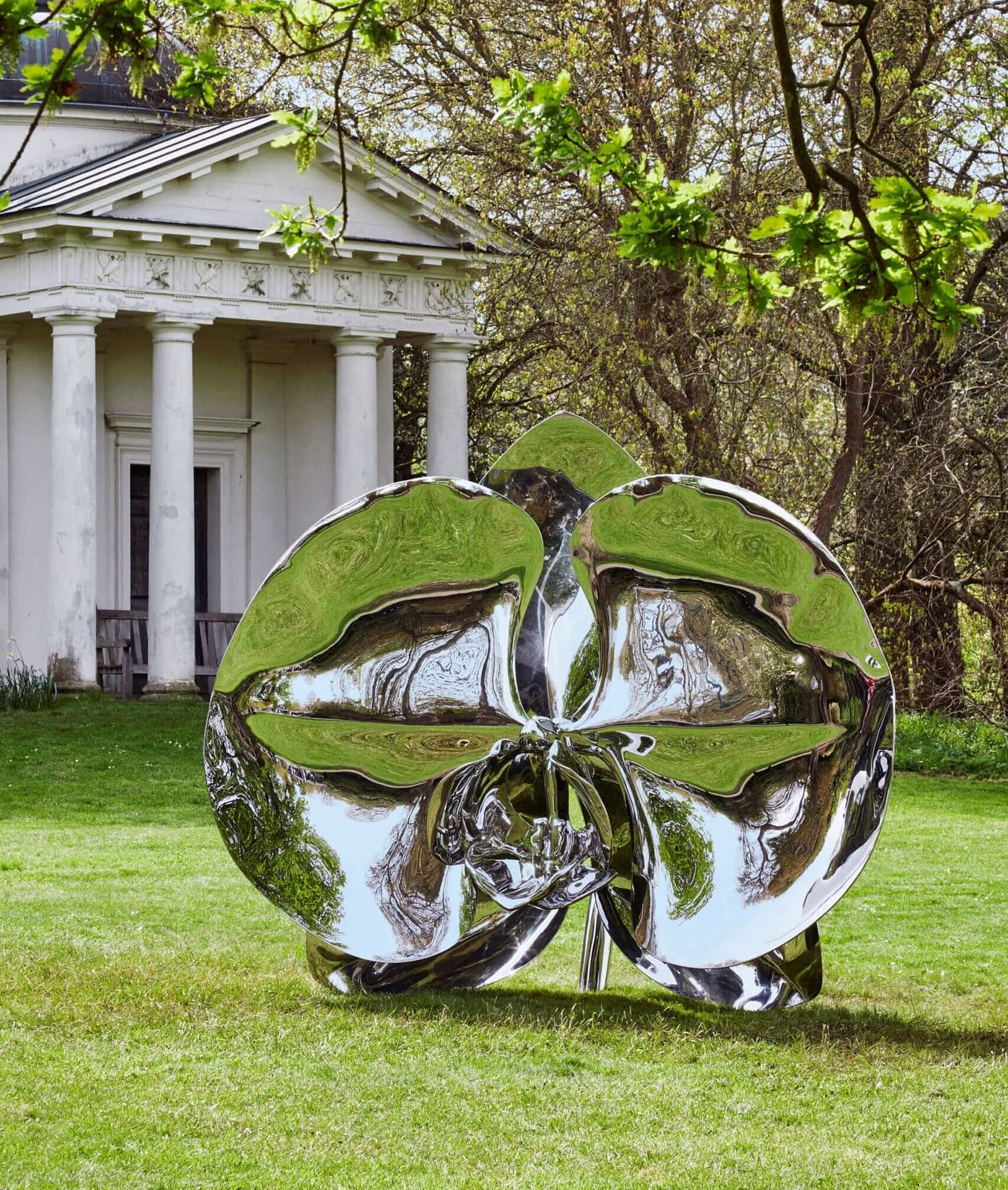 a larger-than-life chrome-coated bronze sculpture of an orchid on a lawn with trees and a classical building in the background