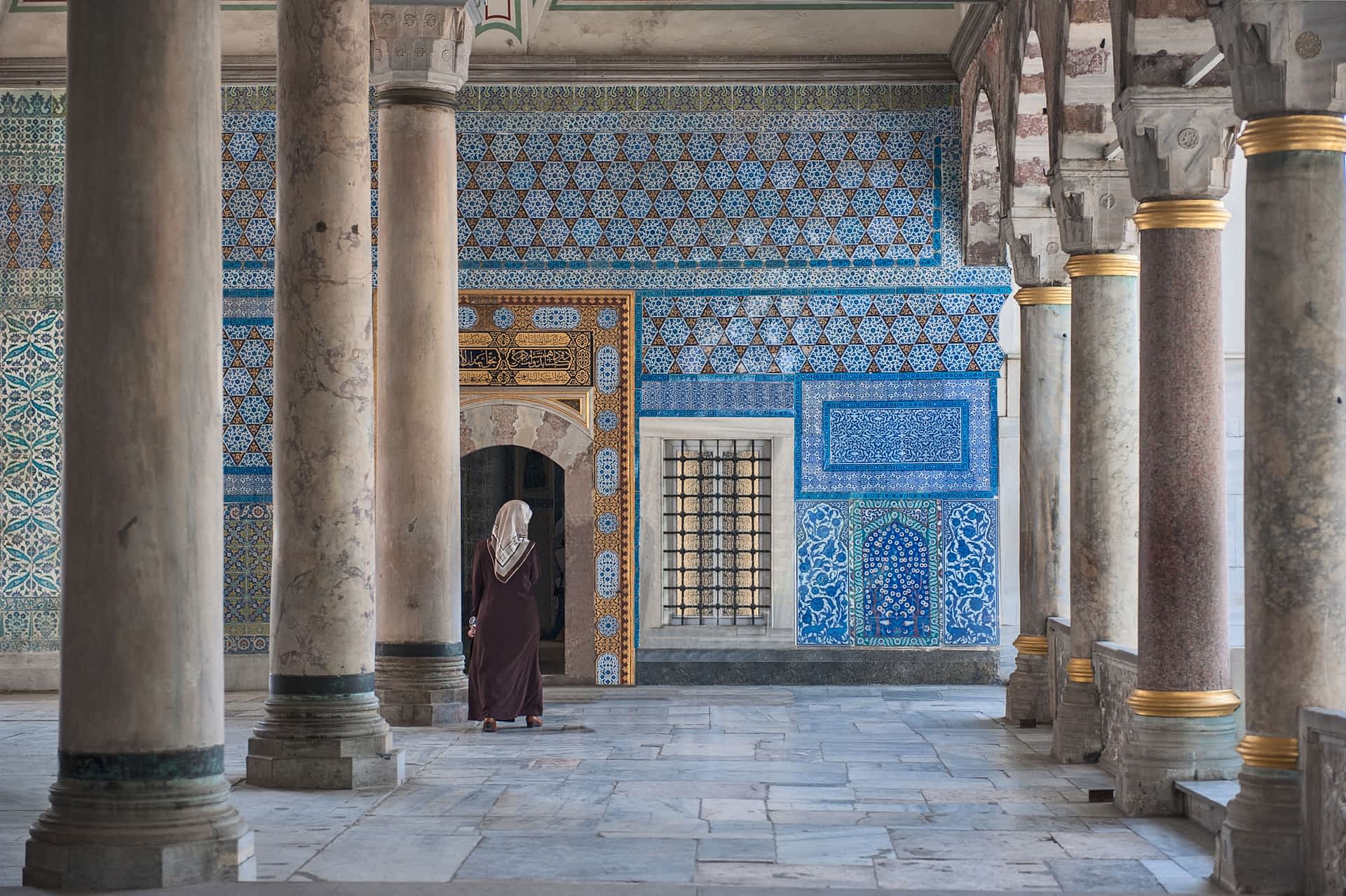 A woman walks towards a doorway in a wall covered with intricately patterned blue tiles at Topkapı Palace. The room has several tall, marble columns.
