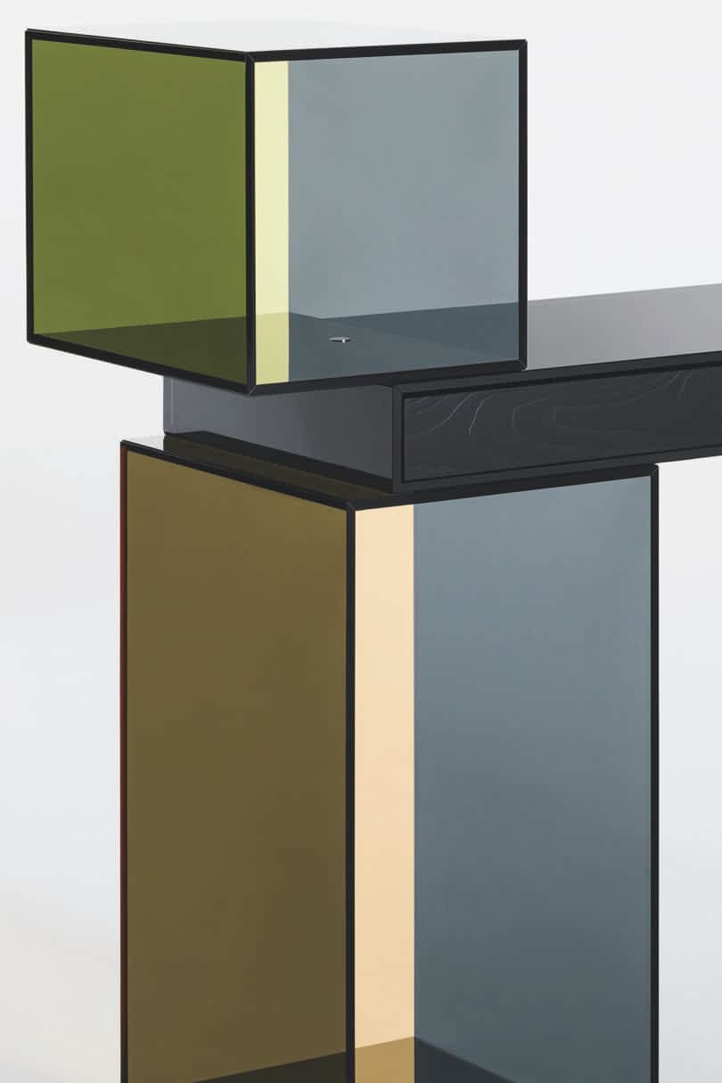 Detail of a modern, minimalist desk with an asymmetrical design featuring black surfaces and transparent colored panels in green and orange. One leg is black while the other supports a green cube.