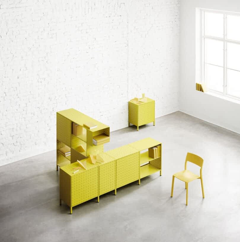 A minimalist workspace featuring yellow perforated String Furniture, including a chair, a cabinet, shelves, and a side table, against a white brick wall and a large window.