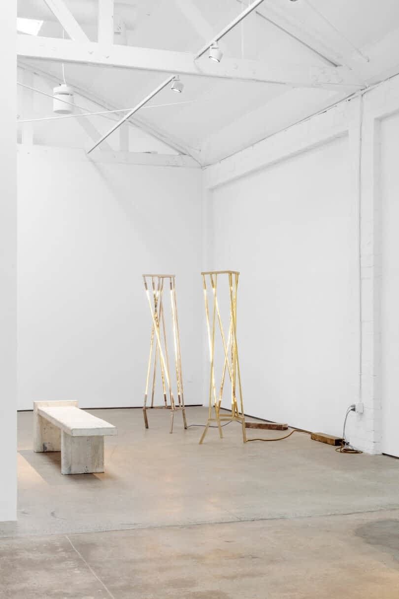 A minimalistic gallery space with a white bench, two wooden sculptures, white walls, and concrete floors. The ceiling has exposed beams and spotlights.