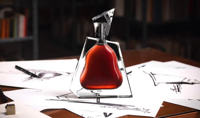 On a desk cluttered with sketches and drawing instruments, a uniquely shaped perfume bottle filled with amber liquid stands beside a Richard Hennessy cognac bottle.
