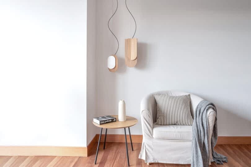 A minimalist living space with a white armchair, striped cushion, small wooden side table holding a vase with branches, and a book. Two wooden pendant lights hang on the white wall