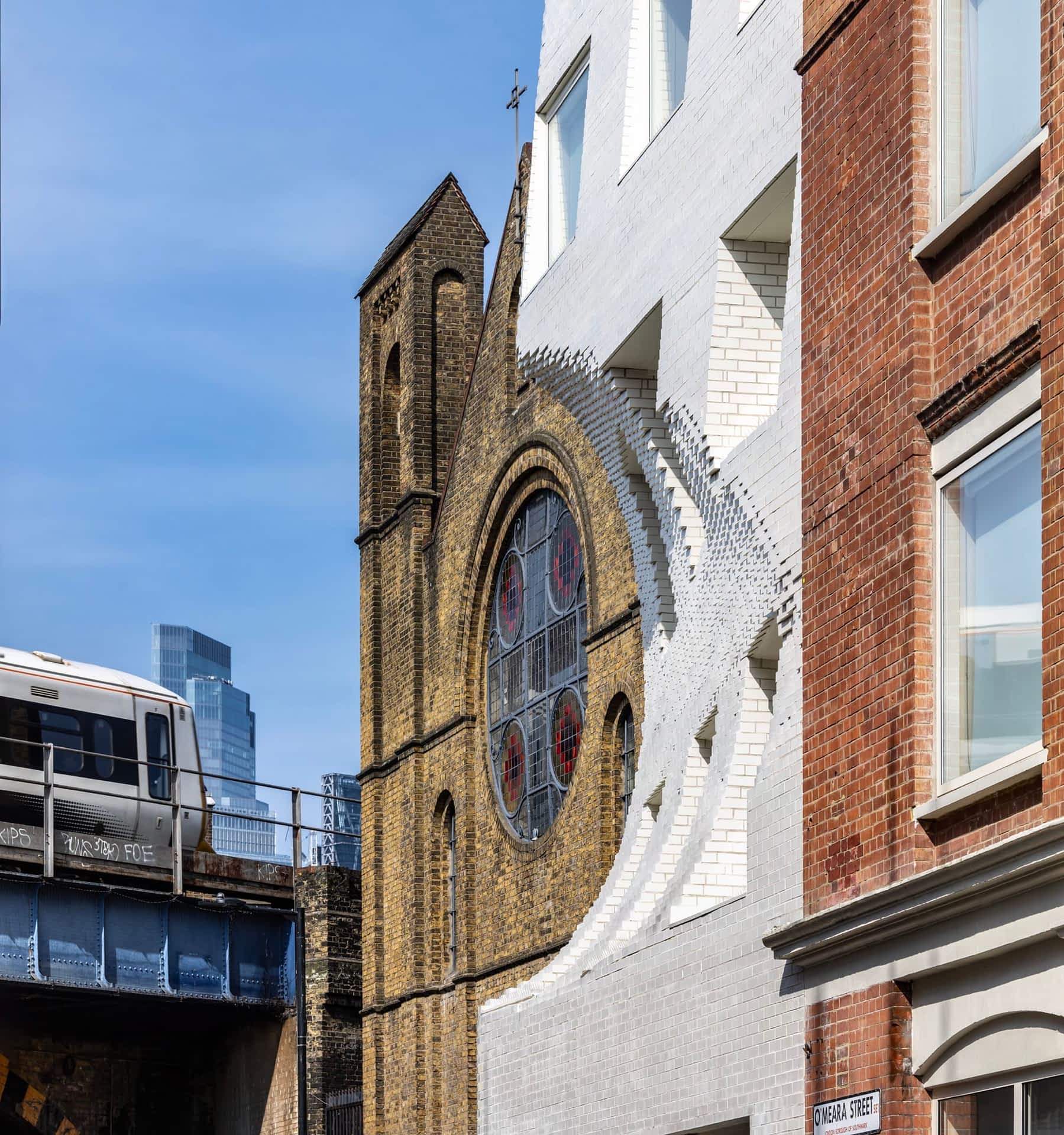 a white brick facade in London looks like it has a large scoop carved out of it, revealing a perfectly framed circular window on an adjacent curch