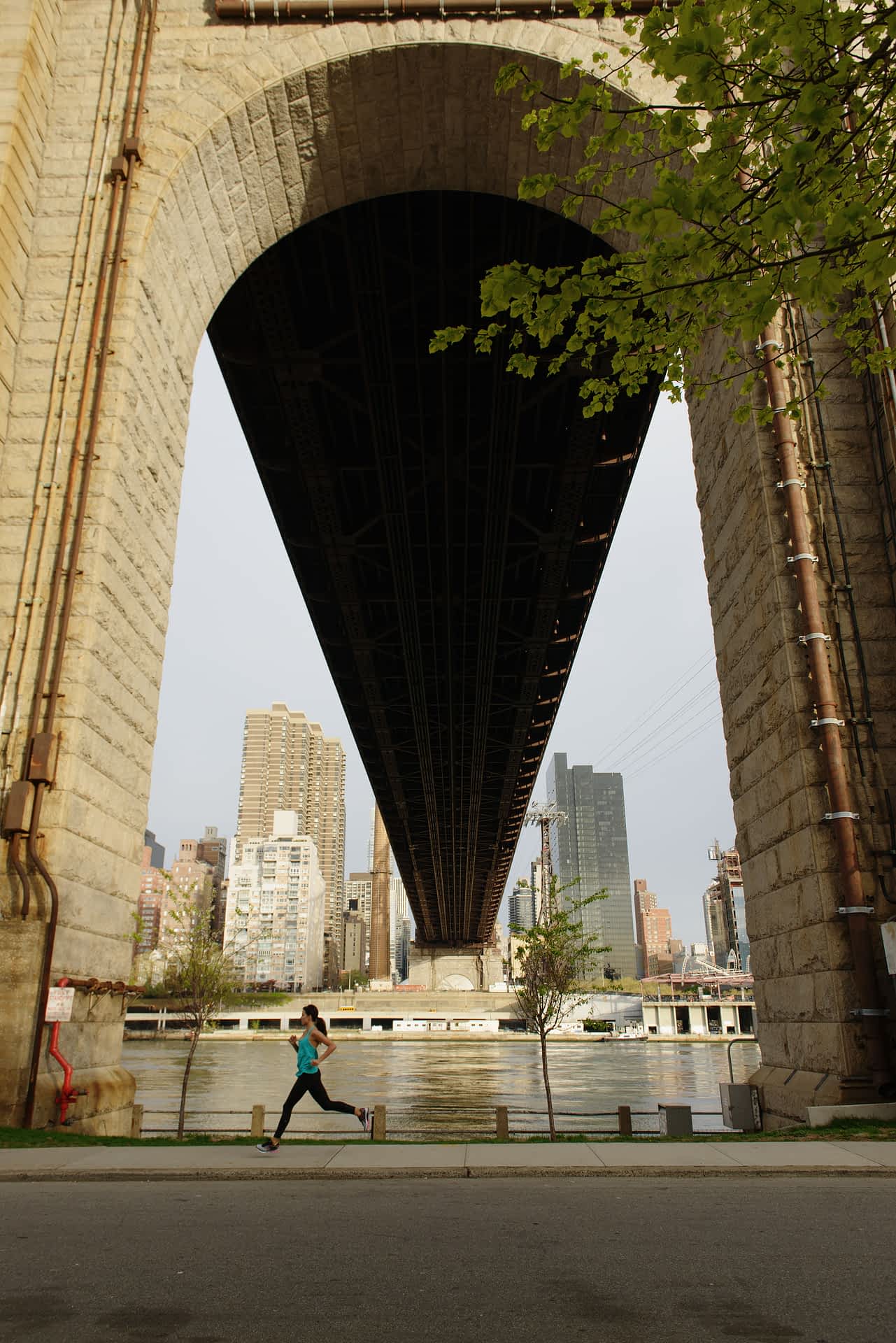 Roosevelt-Island-New-York.jpg A woman is running on the sidewalk under one of the arches of the Queensboro Bridge on Roosevelt Island, New York. Across the river, skyscrapers and large buildings can be seen.