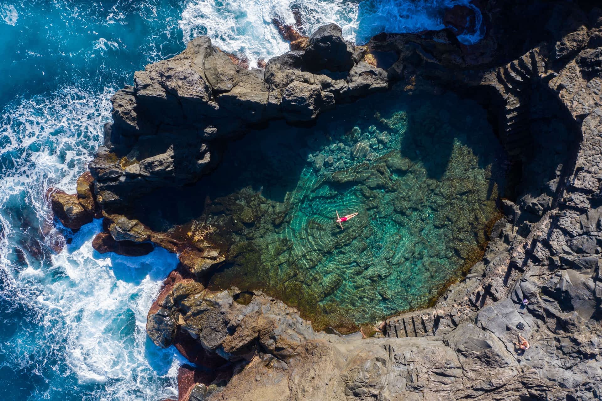 Tenerife A woman in a red bathing suit floats in a large rock pool next to the crashing surf
