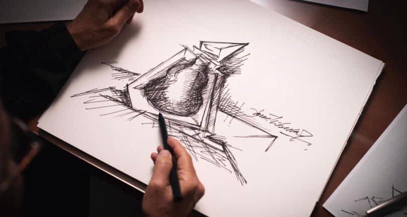 A person sketches an abstract design of a cognac bottle on white paper using a pen, their hand, holding the pen, in the foreground.