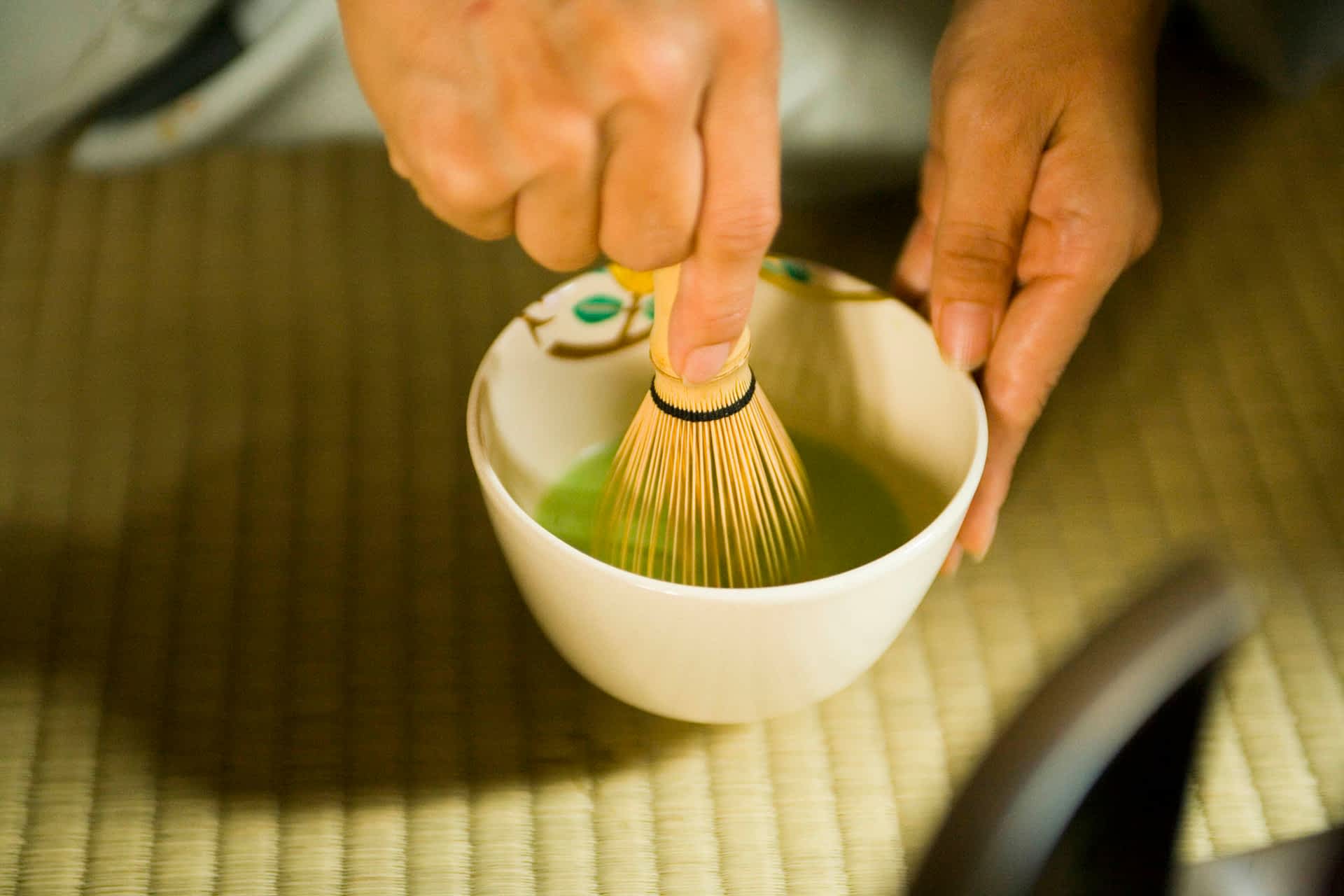 A hand with a whisk stirs matcha -- powdered green tea -- in a small white bowl on a woven mat