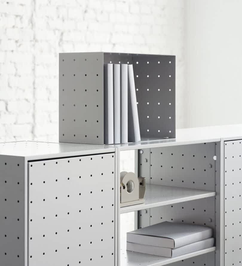 A minimalist shelving unit from the String Furniture X FUWL Center collaboration, featuring a perforated metal design and holding a gray box with books, a tape dispenser, and other office supplies. The unit stands against a white brick wall backdrop, enhancing your workspace furniture.