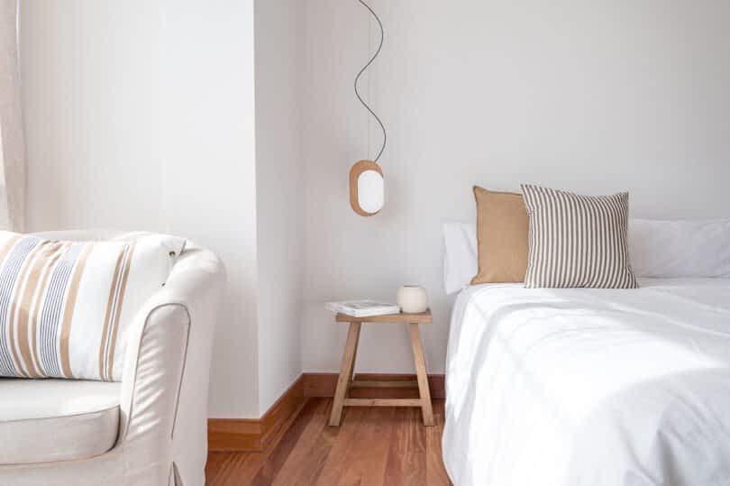 A minimalist bedroom features a white bed with striped pillows, a wooden side table holding a candle and book, and a beige armchair with striped cushions. A pendant light hangs above the table.