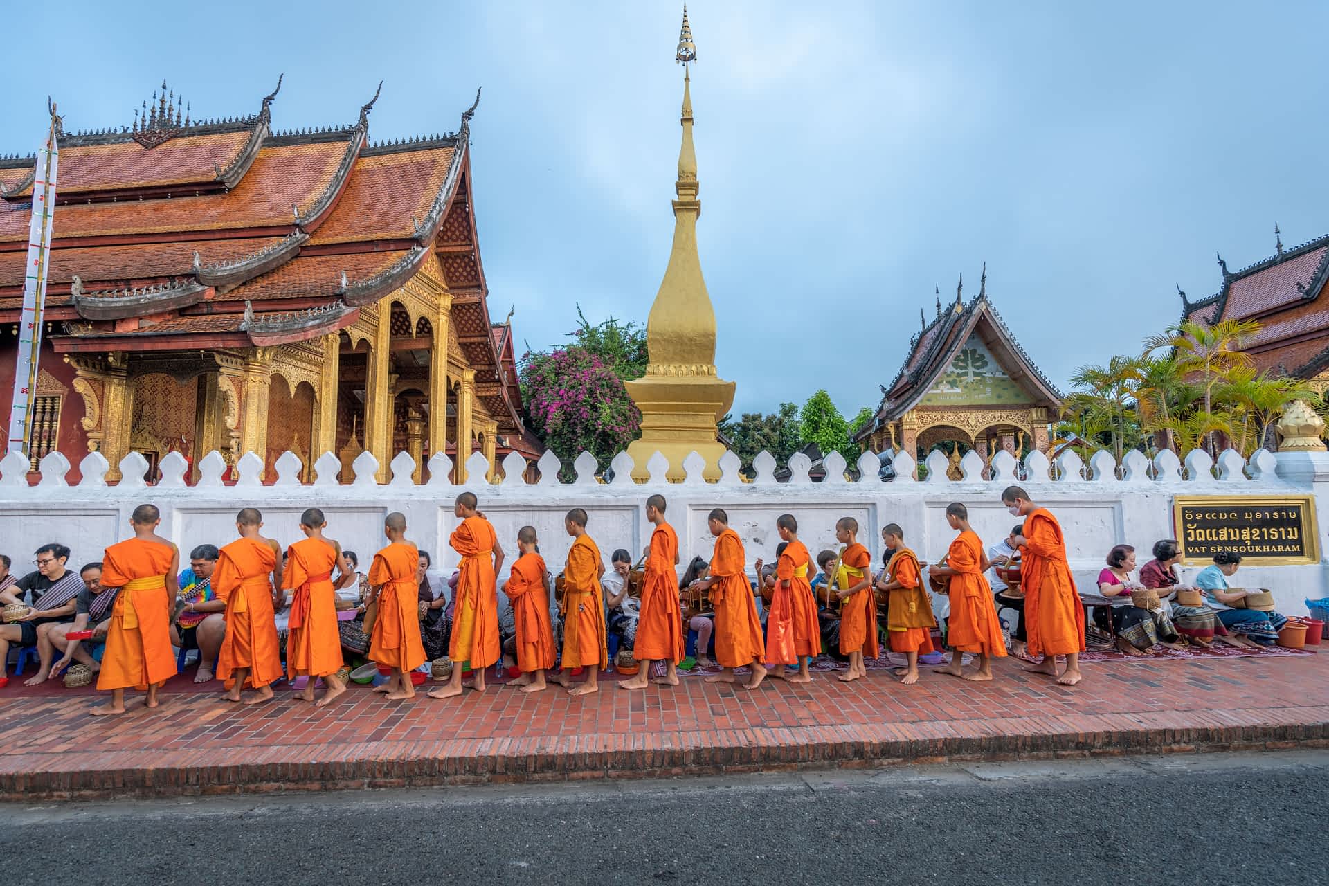shutterstock2193063343.jpg A row of monks in orange robes pass in front of a temple collecting money off people