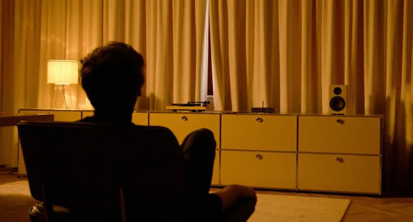 Shadowy silhouette of a man seated listening to his home audio turntable system set against a low console with curtains in the background and warm illuminated light.