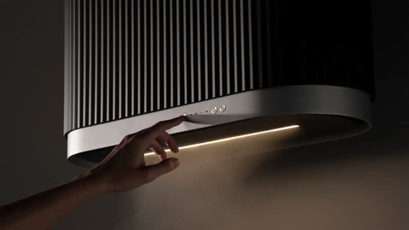 A hand turns on a modern Mono kitchen hood by Ciarko Design with built-in lighting, casting a warm glow on the stove below.