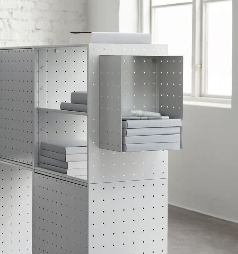 A minimalistic, white, perforated metal shelf unit from String Furniture X FUWL Center is set against a white brick wall, with books neatly arranged on the shelves and a window in the background. This elegant piece makes an ideal addition to any workspace furniture collection.