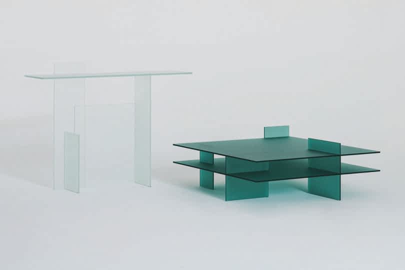 Two minimalist glass tables, one taller and clear, the other shorter and green, displayed against a plain background.