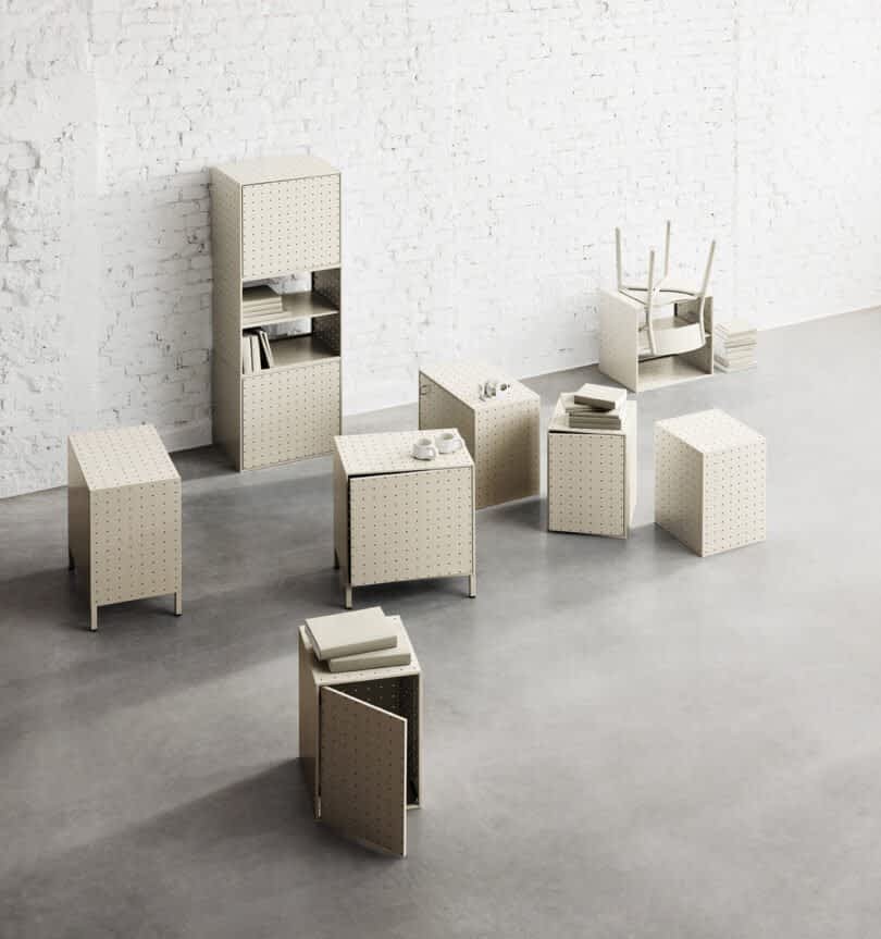 A minimalist room with white brick walls features various beige modular String Furniture pieces, including tables, chairs, and storage units, all arranged on a gray concrete floor.