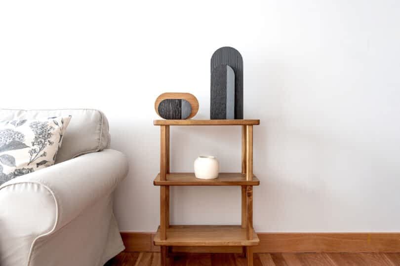 A wooden shelf stands against a white wall, holding a white candle and two wooden lamps. A beige sofa with a patterned cushion is positioned to the left of the shelf.