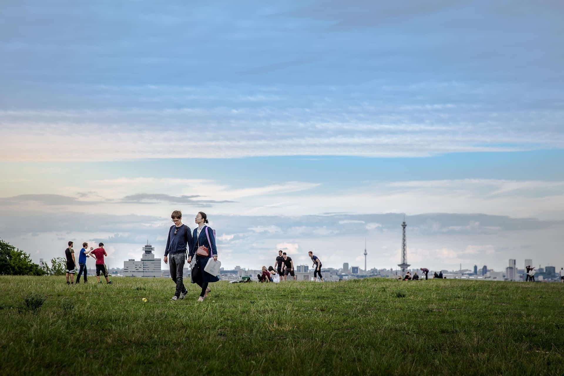 GettyImages-949380682.jpeg Two people walk hand in hand through a park as others play a game behind them. The cityscape rises in the distance