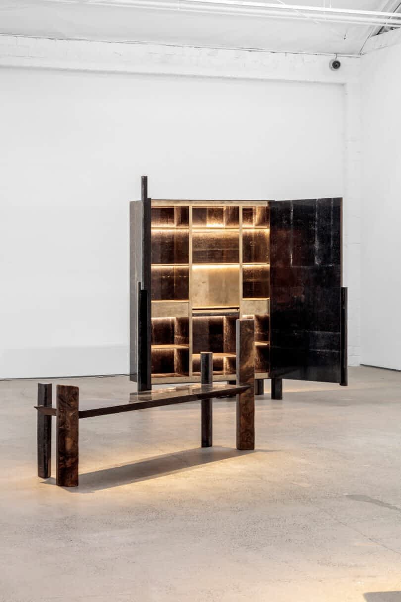 A modern art installation by Vincenzo De Cotiis features a minimalist bench and a tall, illuminated shelving unit with open and closed compartments in a white-walled gallery.