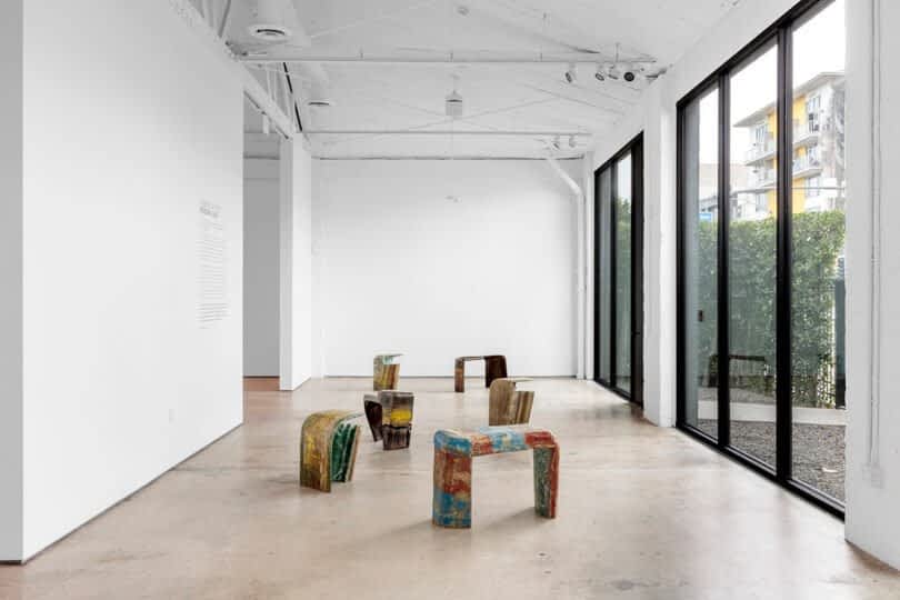 Modern art gallery featuring colorful, abstract sculptures made from various materials, displayed on a polished concrete floor. Large windows line the right wall, letting in natural light. The gallery offers an elegant yet raw aesthetic that captivates visitors.