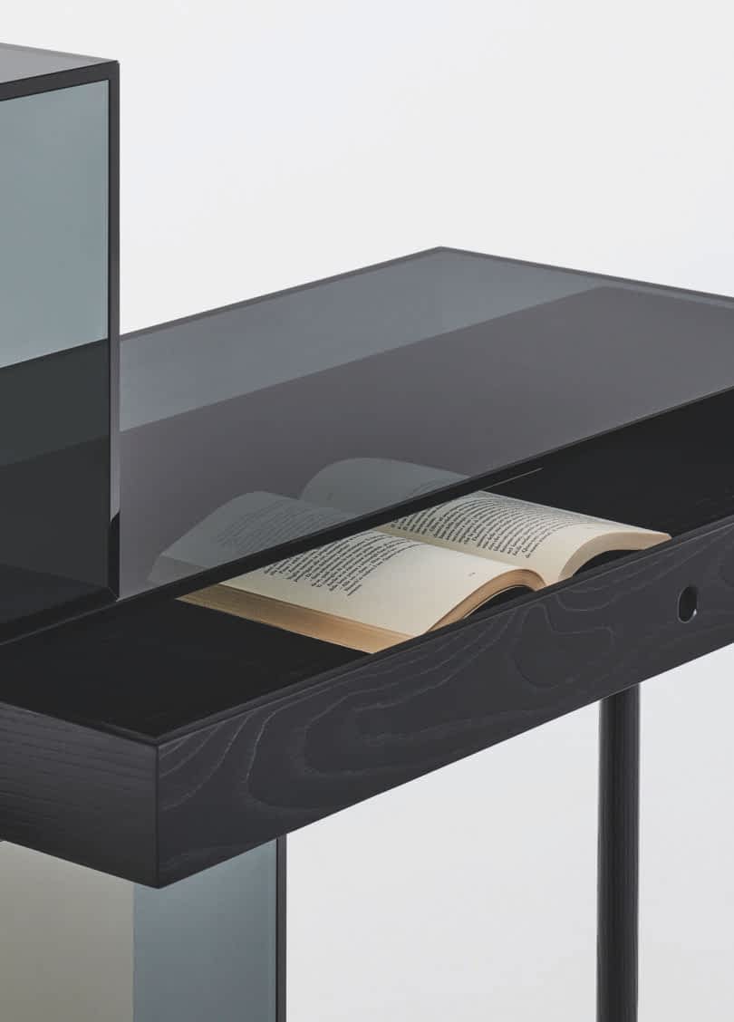 Detail of a modern, minimalist desk with an asymmetrical design featuring black surfaces and transparent colored panels in green and orange. One leg is black while the other supports a green cube.