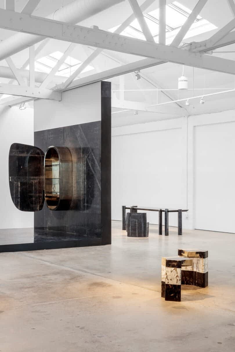 Modern art installation in a gallery featuring geometric black sculptures and furniture, with high ceilings and exposed white beams.