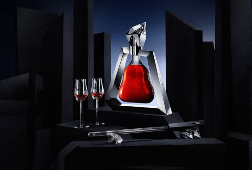 A Richard Hennessy Magnum cognac bottle stands proudly on a stand, its triangular decanter filled with red liquid. Flanked by two filled glass flutes and a crystal-stoppered rod, it is set against a backdrop of geometric black shapes.