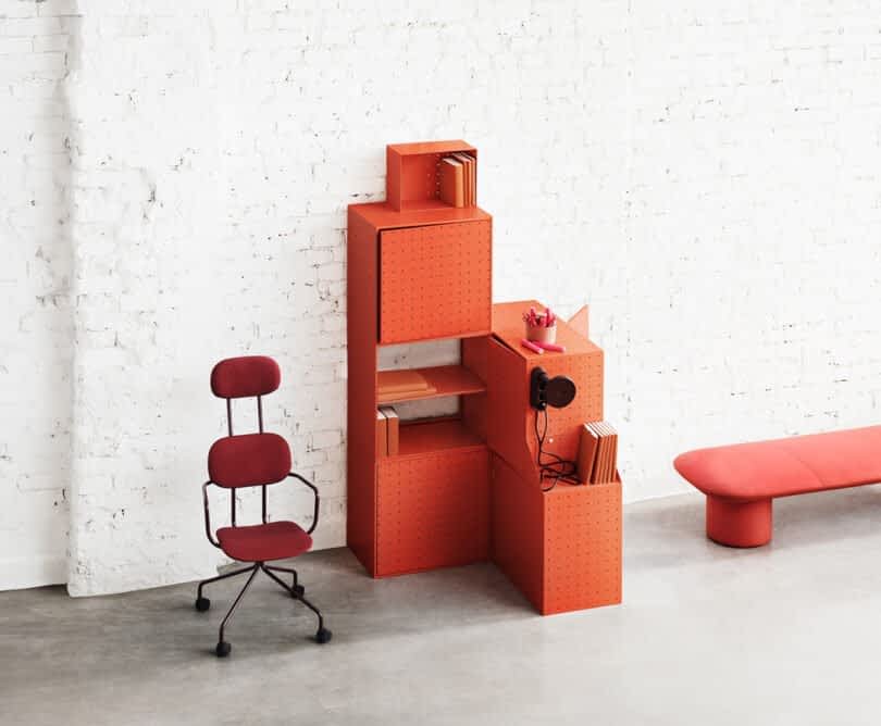 A modular orange storage unit by String Furniture X FUWL Center stands against a white brick wall, filled with books and accessories. A red cushioned chair and a matching bench are positioned nearby on the concrete floor, making for an efficient workspace furniture arrangement.