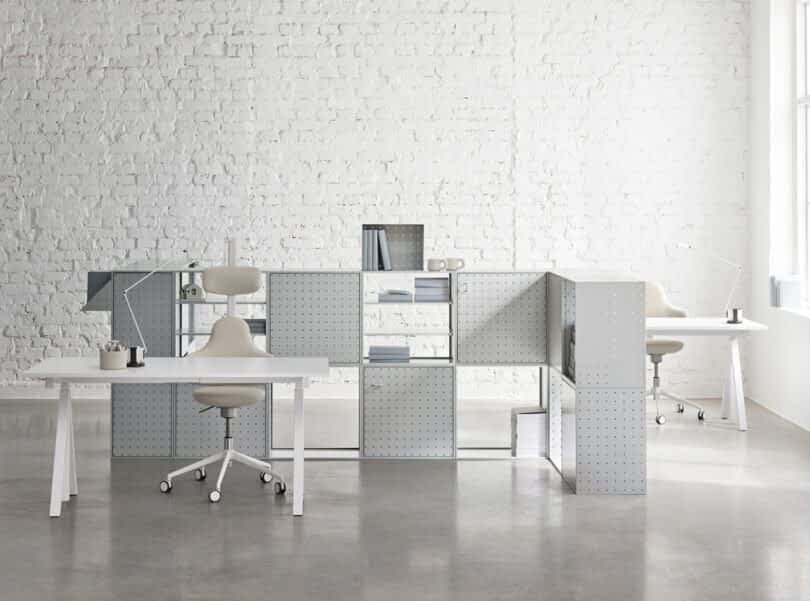 Modern office space with String Furniture X FUWL Center workspace furniture, featuring two white desks, white chairs, and silver shelving units against a white brick wall. Shelves hold various office supplies and books. Natural light enters through a nearby window.
