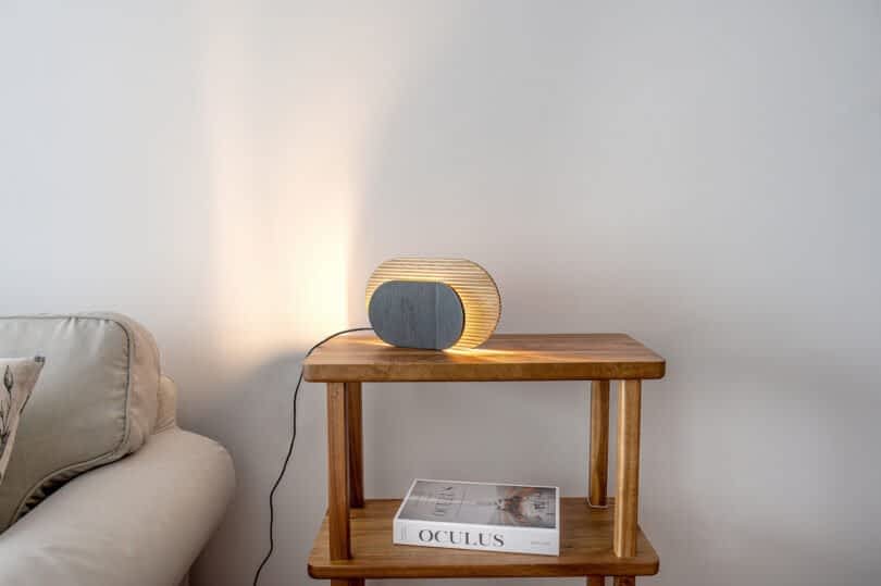 A wooden side table with decorative objects: a modern, oval-shaped wooden and black lamp, and a small, light-colored ceramic vase. Part of a light gray couch is visible on the left.