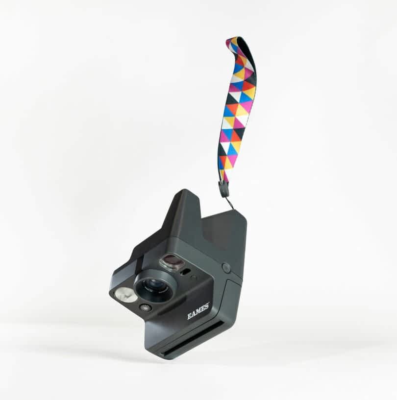 A black instant camera with a geometric-patterned strap is suspended in mid-air against a plain white background.