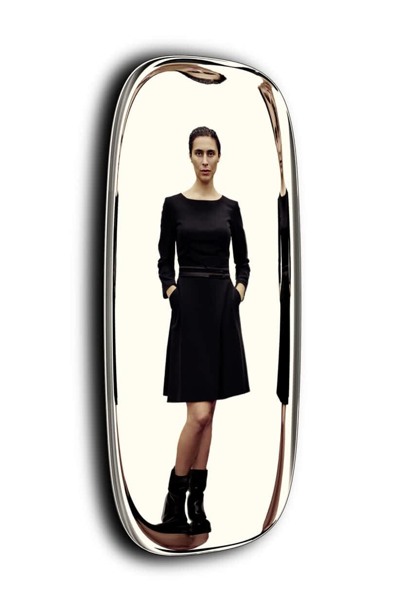 A woman in a black dress and ankle boots stands reflected in a tall metallic framed mirror.
