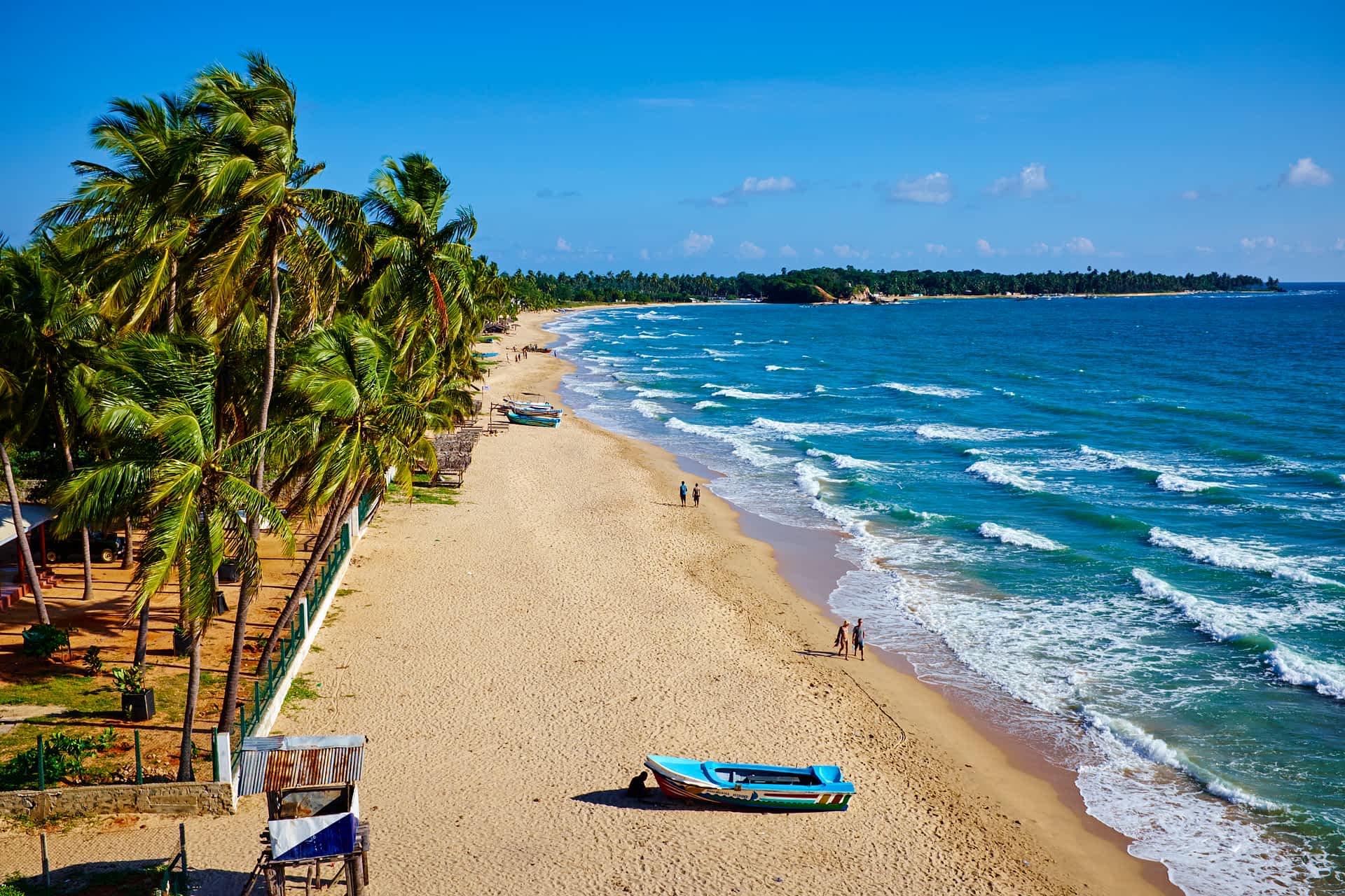 Uppuveli beach, Trincomalee, Sri Lanka A scenic aerial shot of a mostly empty beach with palm trees, golden sand and gentle turquoise waves, two couples walking by the water and a small boat on the sand in the foreground